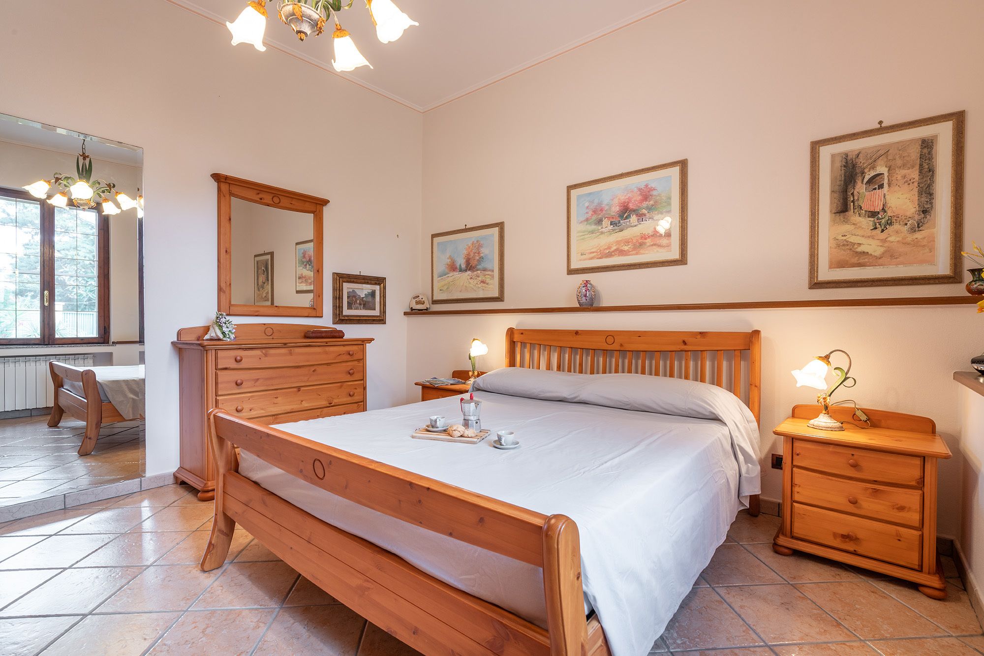 Een slaapkamer met een houten bed, een dressoir, een nachtkastje en een spiegel. Drie ingekaderde foto's hangen aan de muur.