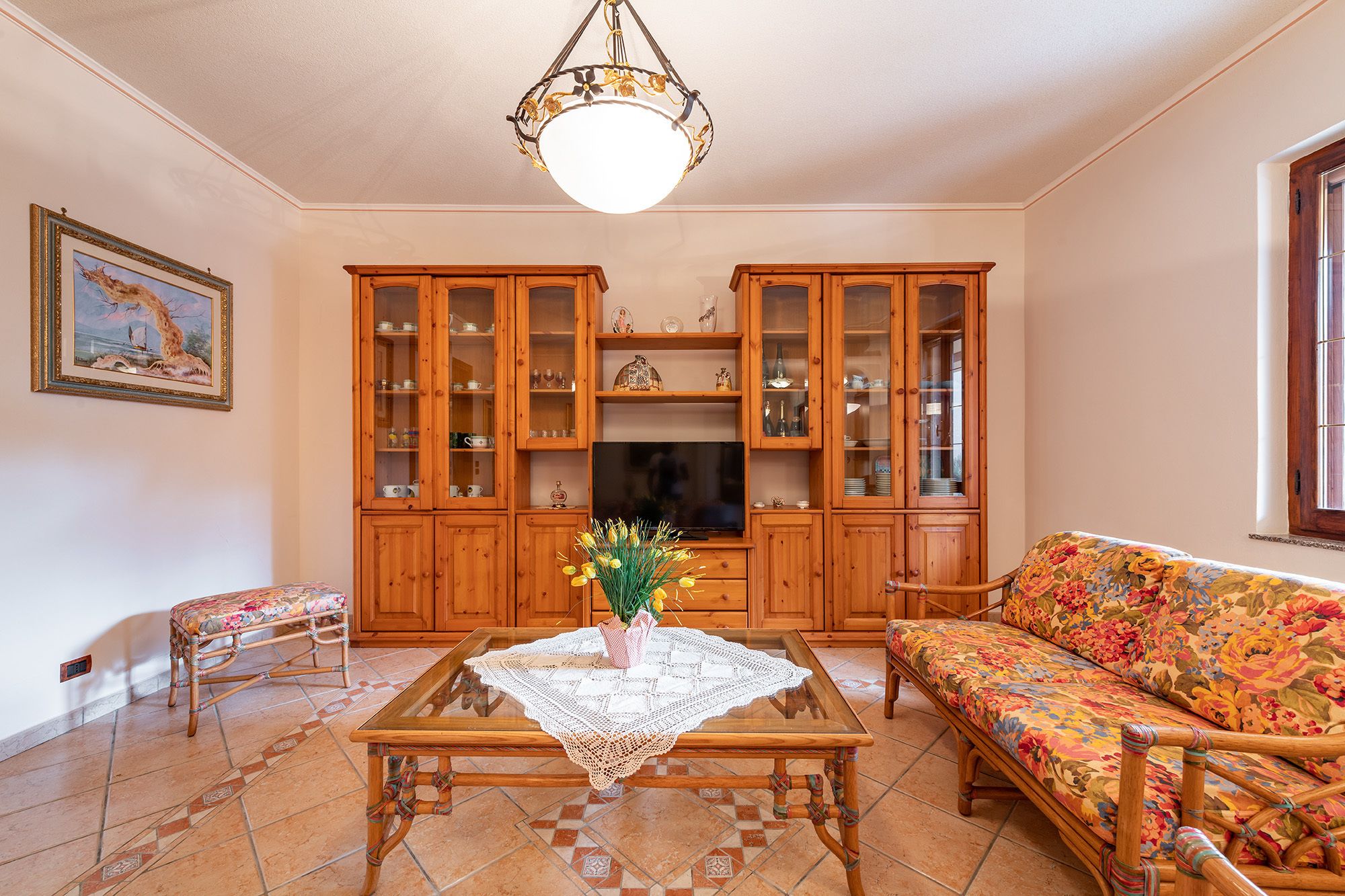 Una sala de estar con una mesa de centro de vidrio, un sofá floral, armarios de madera y un televisor.
