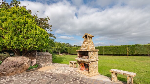 A stone outdoor barbecue area with a stone chimney, surrounded by a stone pathway and benches. - AffittaSardegna - Villetta Tennis 1 (Alghero)