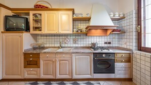 A kitchen with wooden cabinets, a built-in oven, a stove, and a television on the countertop. - AffittaSardegna - Villetta Tennis 1 (Alghero)