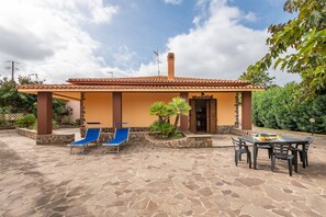 A house with a tiled roof, a covered patio with blue lounge chairs, and a stone patio with a table and chairs.