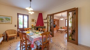 A dining area with a table set for four, surrounded by wooden chairs, a red fireplace, and a view into a living room with a sofa and a television. - AffittaSardegna - Villetta Tennis 1 (Alghero)