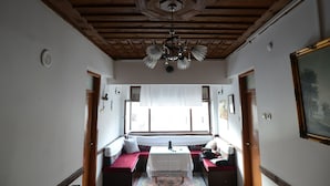 A room with a wooden ceiling, a chandelier, a bench with cushions, a table with a white tablecloth, and a window with a view. - Huseyin Bey Konagi (Safranbolu)