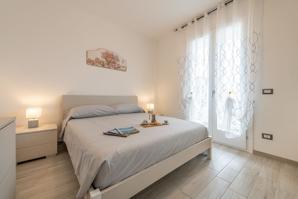 Ein Schlafzimmer mit einem Bett, Nachttischen, einer Fensterbank und einer Wandlampe, sowie einem Fenster mit gemusterten Vorhängen.