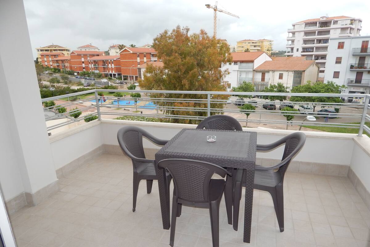 Ein Balkon mit Tisch und Stühlen, der auf einen Wohnbereich mit Gebäuden und einem Schwimmbad blickt.