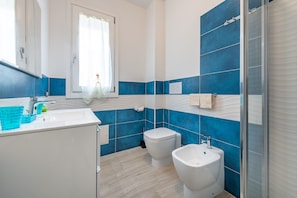 Un baño con lavamanos blanco, dos inodoros, cabina de ducha con mampara de vidrio y paredes con azulejos blancos y azules.