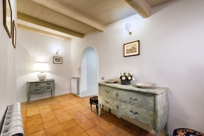 A room with a tiled floor, a wooden ceiling, a dresser with drawers, a small table with a lamp, and a radiator.