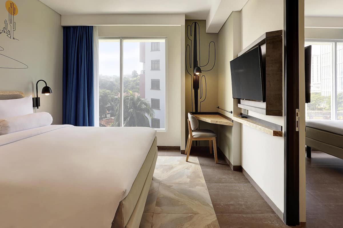 Une chambre d’hôtel équipée d’un lit, d’un bureau, d’une chaise, d’une télévision et d’une grande fenêtre donnant sur des immeubles.