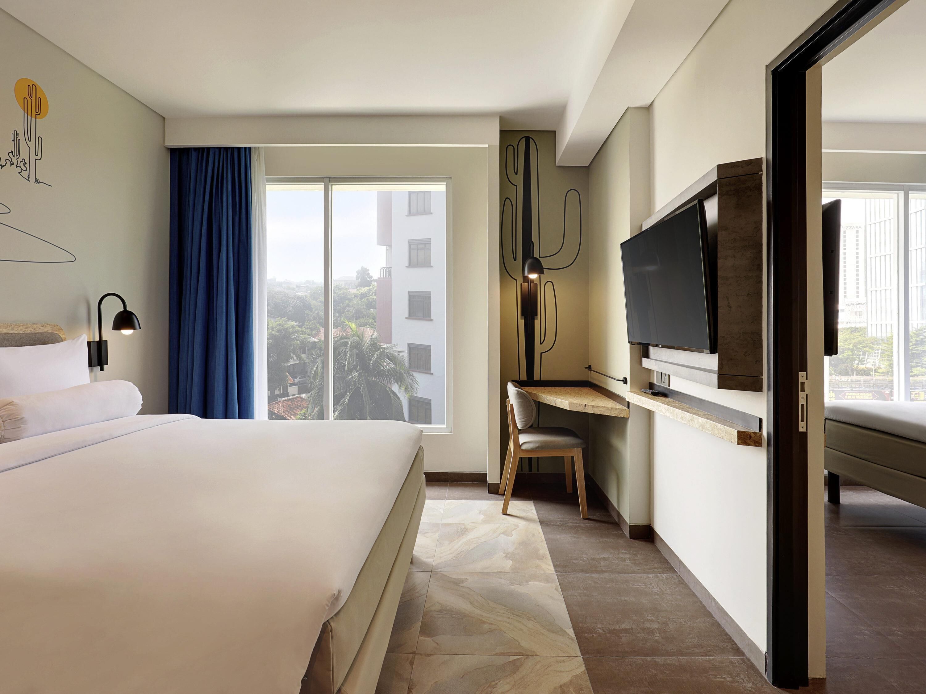 Ein Hotelzimmer mit Bett, Schreibtisch, Stuhl, Fernseher und einem großen Fenster mit Blick auf Gebäude.