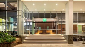 Un moderno vestíbulo de hotel con fachada de vidrio, un mostrador de recepción y una escalera que conduce a los niveles superiores.
