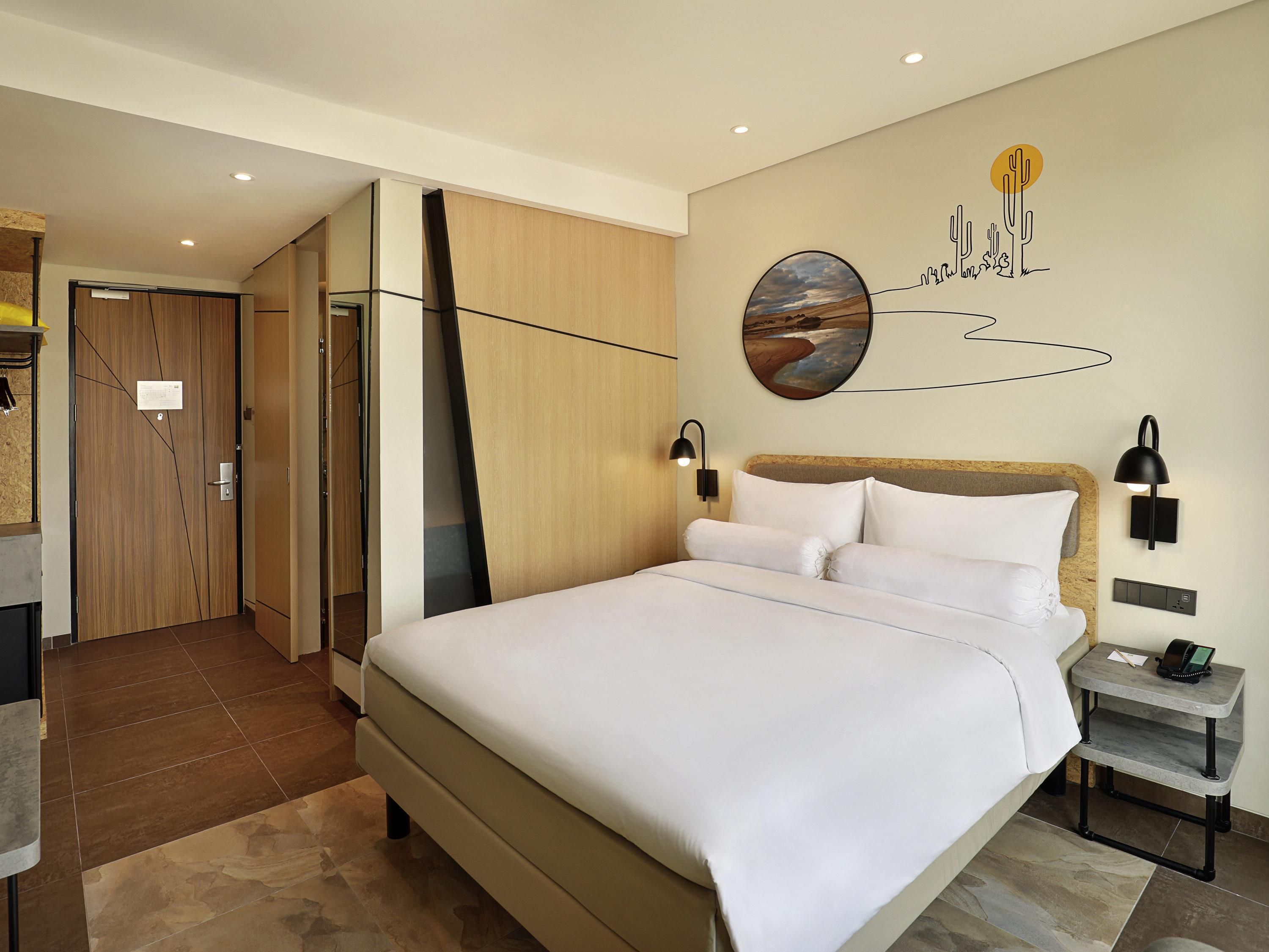 Camera d'albergo moderna con un letto grande, un comodino, una lampada a parete e un dipinto di un paesaggio desertico.