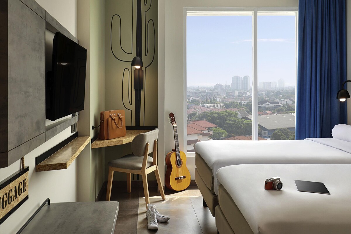 Una camera d'albergo con un letto, una scrivania con una sedia, una chitarra, una televisione e vista sulla città.