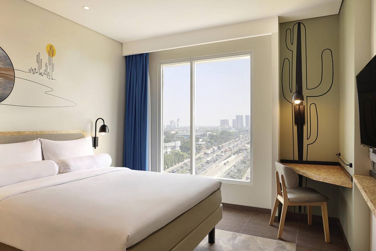 Hotelkamer met een groot bed, een bureau, een stoel, een televisie, een raam met uitzicht over de stad en een kunstwerk aan de muur.