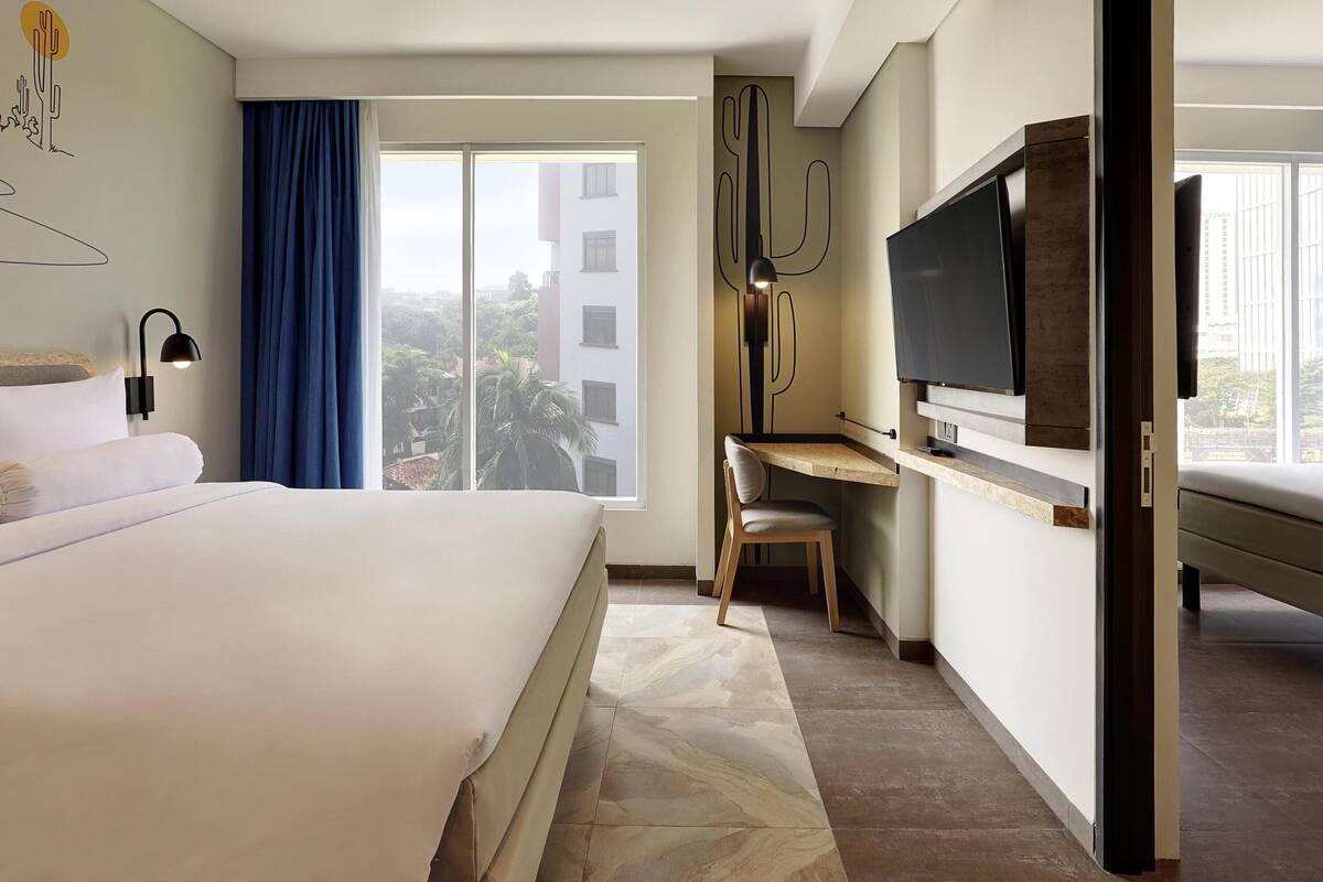 Een hotelkamer met een bed, een bureau, een stoel, een televisie en een groot raam met uitzicht op gebouwen.