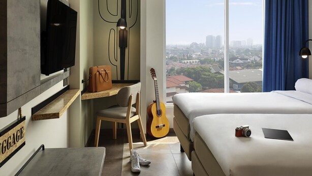 A hotel room with a bed, a desk with a chair, a guitar, a television, and a view of the city. - ibis Styles Jakarta Simatupang (Jakarta)