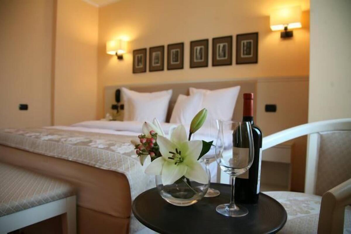 Une chambre d’hôtel avec un lit soigneusement fait, une table de chevet sur laquelle se trouvent une bouteille de vin et un verre, et un vase rempli de fleurs.