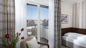 A hotel room with a bed, a chair, a vase of flowers, and a balcony with a view of buildings. - Hotel Harheimer Hof (Frankfurt)
