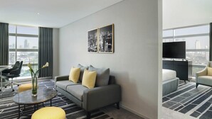 Une chambre d’hôtel moderne avec un canapé gris, des coussins à motifs jaunes, une table basse en verre, un pouf jaune, un téléviseur à écran plat et une vue sur le paysage urbain à travers de grandes fenêtres.