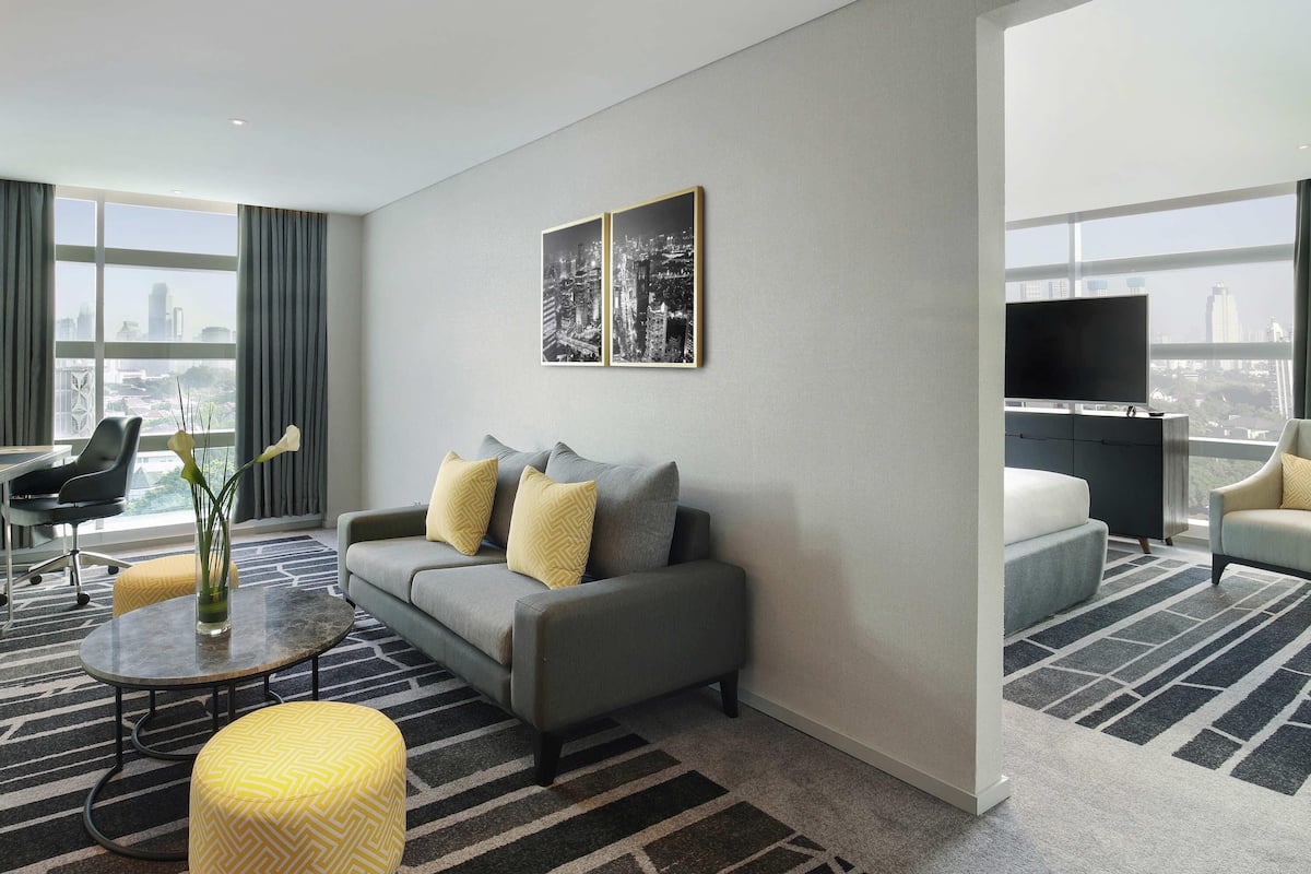 Ein modernes Hotelzimmer mit grauem Sofa, gelben Kissen mit Muster, einem Glastisch, einem gelben Hocker, einem Flachbildfernseher und Blick auf die Stadtlandschaft durch große Fenster.