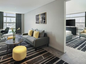 Habitación de hotel moderna con sofá gris, cojines estampados amarillos, una mesa de centro de vidrio, un reposapiés amarillo, un televisor de pantalla plana y vistas a la ciudad a través de amplias ventanas.