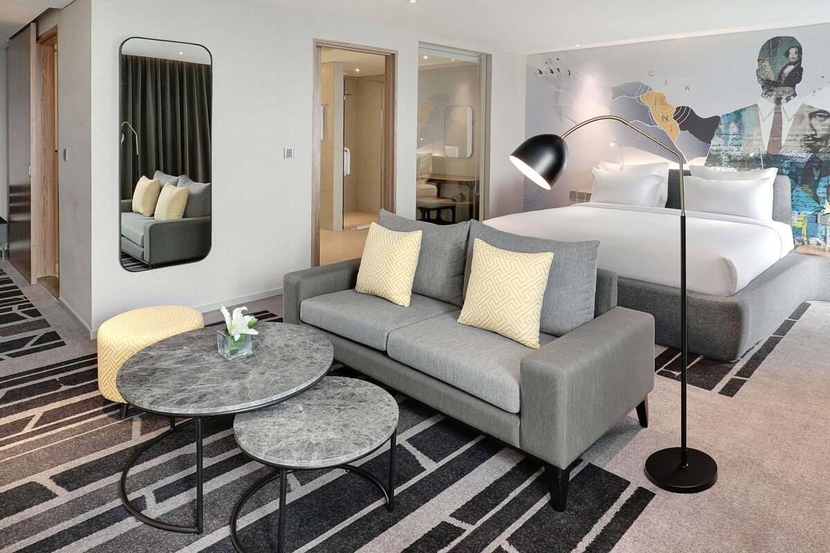 Ein modernes Hotelzimmer mit grauem Sofa, einem runden Couchtisch, einem Bett mit weißer Bettwäsche, einem großen Spiegel und einem Wandgemälde.