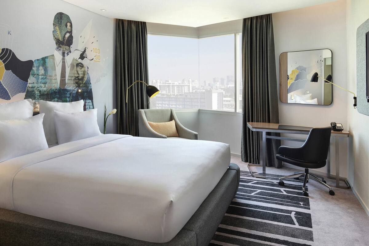 Ein Hotelzimmer mit einem großen Bett, einem Schreibtisch, einem Sessel und einem Spiegel, mit Blick auf die Stadt.