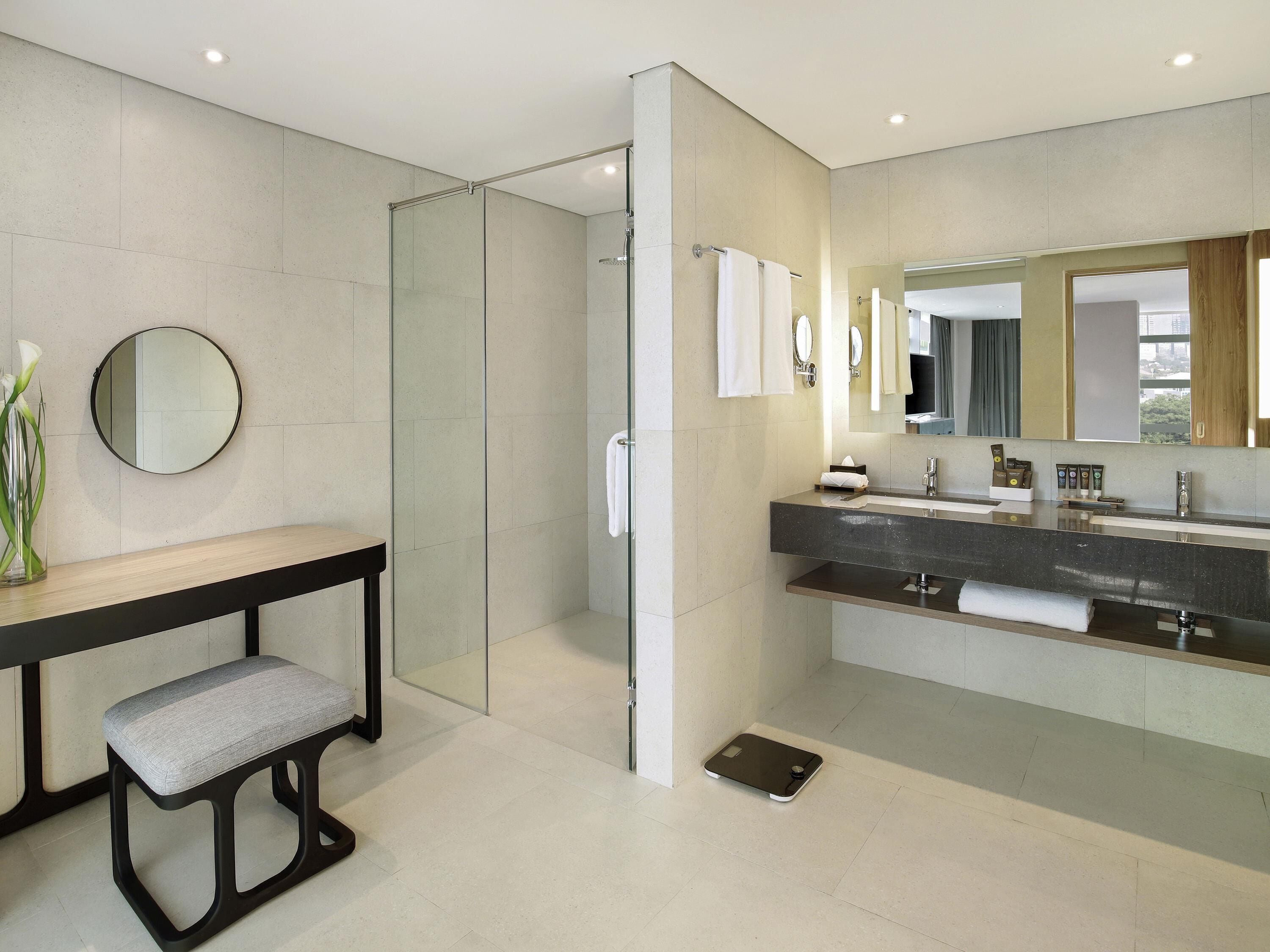 A modern bathroom with a glass shower enclosure, a vanity with a dark countertop, a round mirror, and a bench.