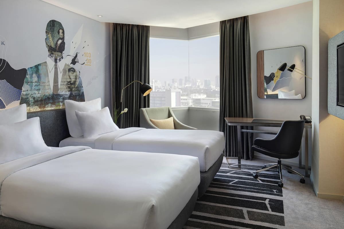 Ein Hotelzimmer mit zwei Betten, einem Schreibtisch, einem Stuhl, einem großen Fenster mit Blick auf die Stadt und Kunstwerken an den Wänden.