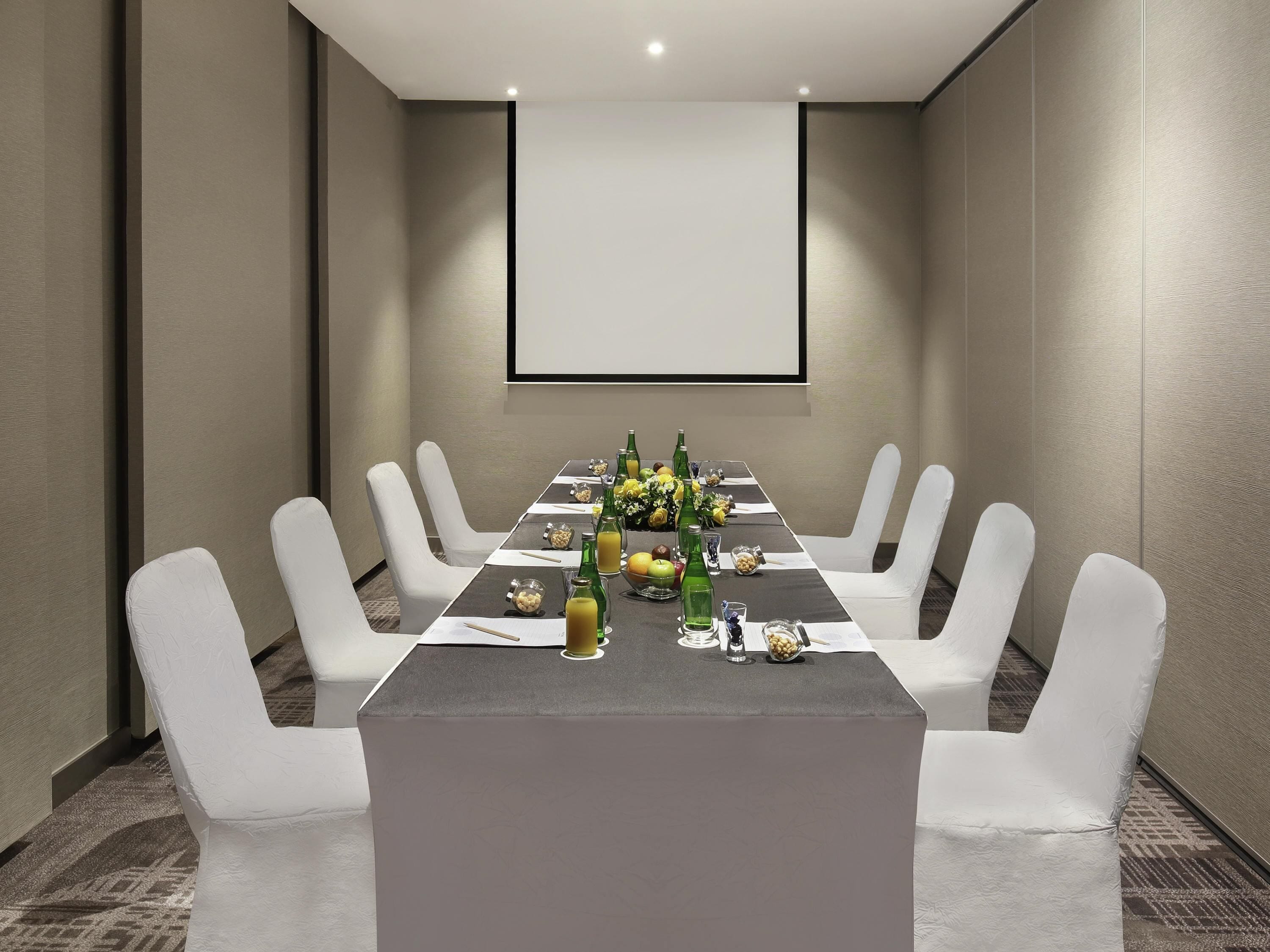 A conference room with a long table, white chairs, and a projector screen.