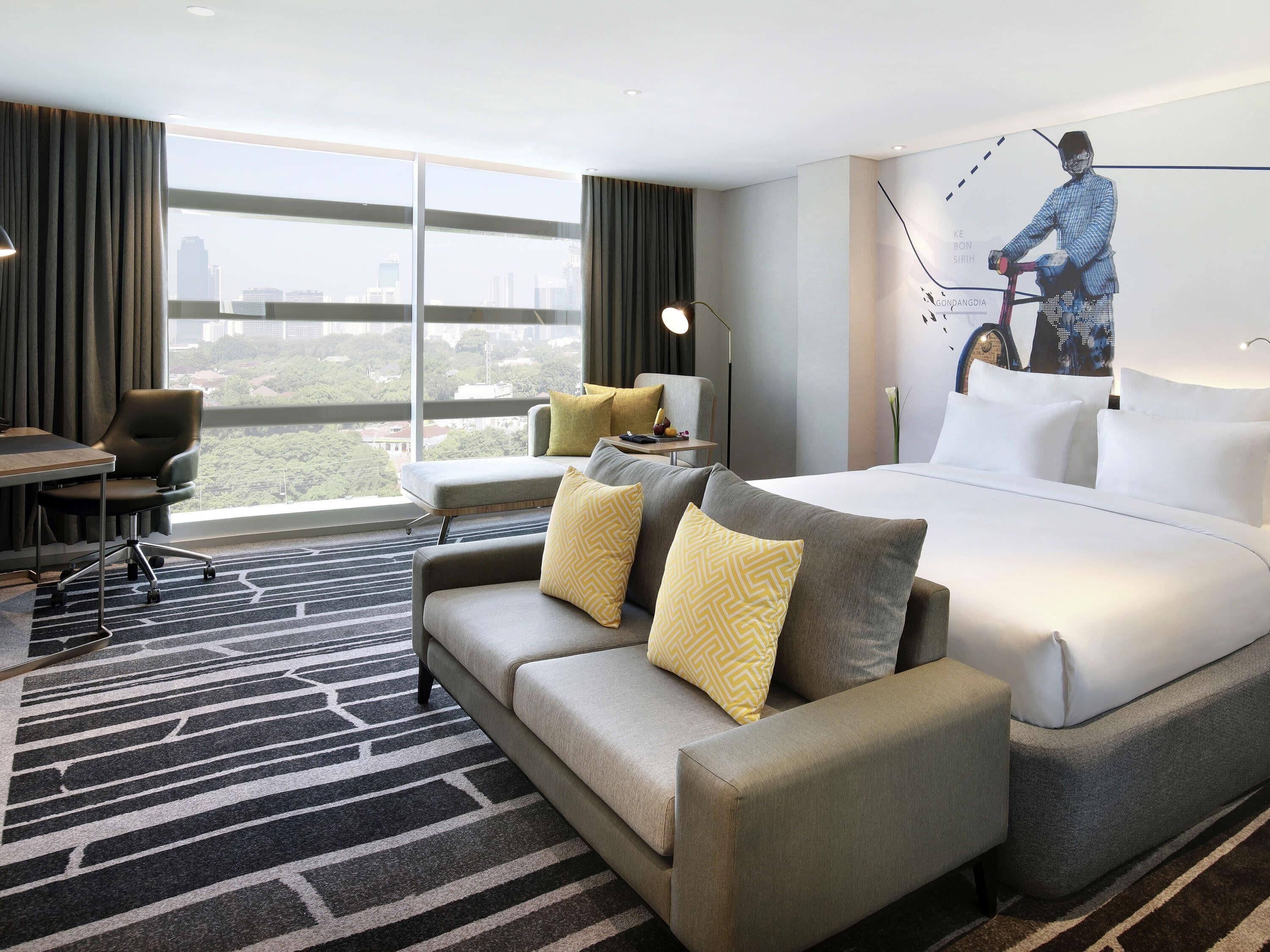 A hotel room with a large window, a sofa, a bed, a desk, and a chair.