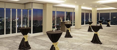 Uma sala de conferências espaçosa com grandes janelas que oferecem vista para a cidade, várias mesas com toalhas pretas e fitas amarelas, e um espaço de bar.