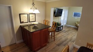 A kitchen with a wooden island, two chairs, a small table, and a TV mounted on the wall. - The Sand Castle (Satellite Beach)
