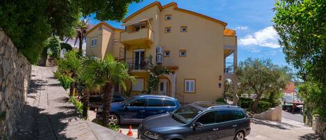A two-story building with a yellow facade, a balcony on the second floor, and parked cars in front.