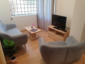 A living room with two grey sofas, a wooden coffee table with a book and a small plant, a TV on a wooden stand, and a window with curtains. - Captain's Club (Rab)