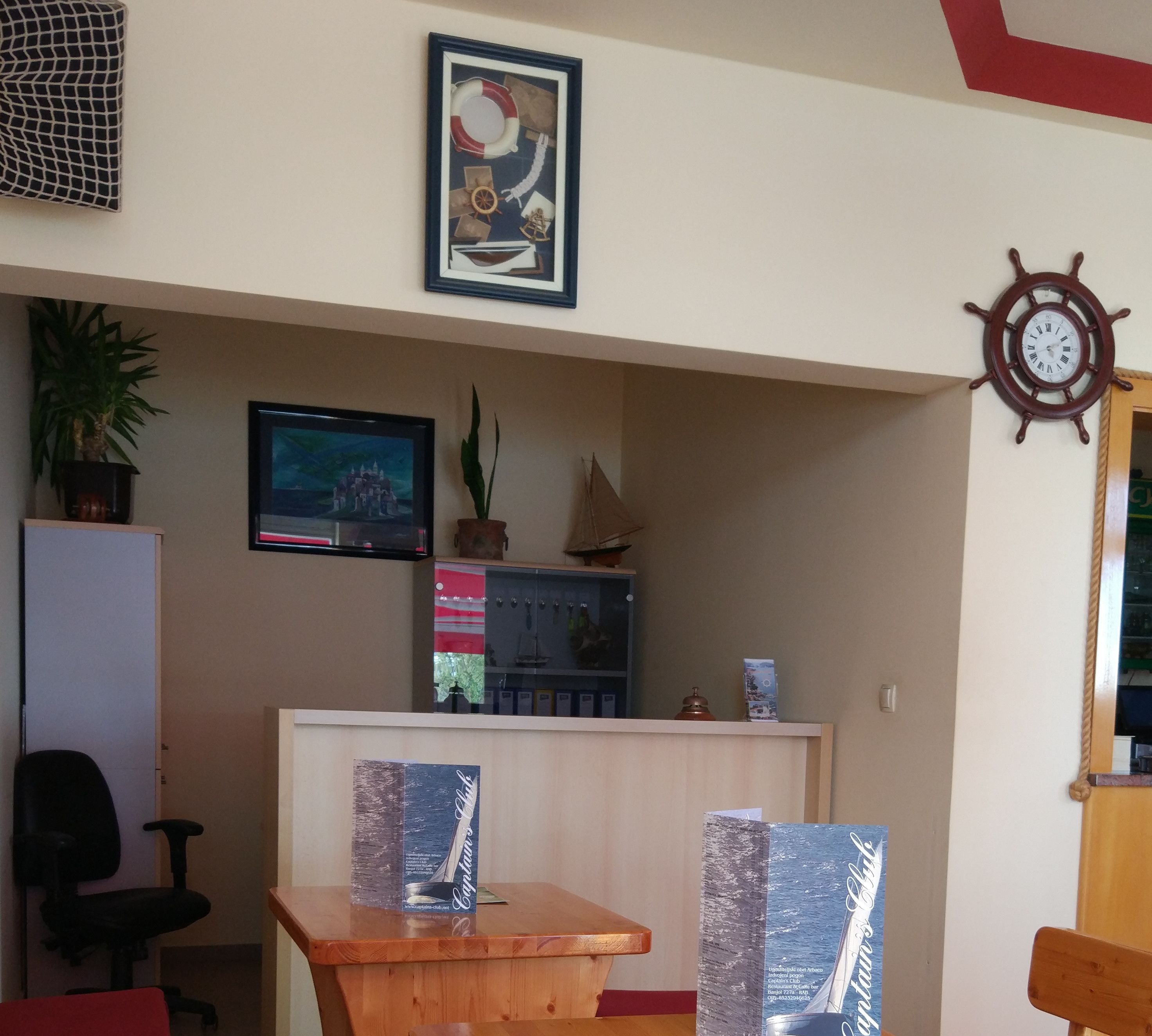 Un'area reception con una scrivania in legno, un'opera d'arte a tema nautico in cornice, un modellino di nave e un orologio a forma di timone fissato alla parete.