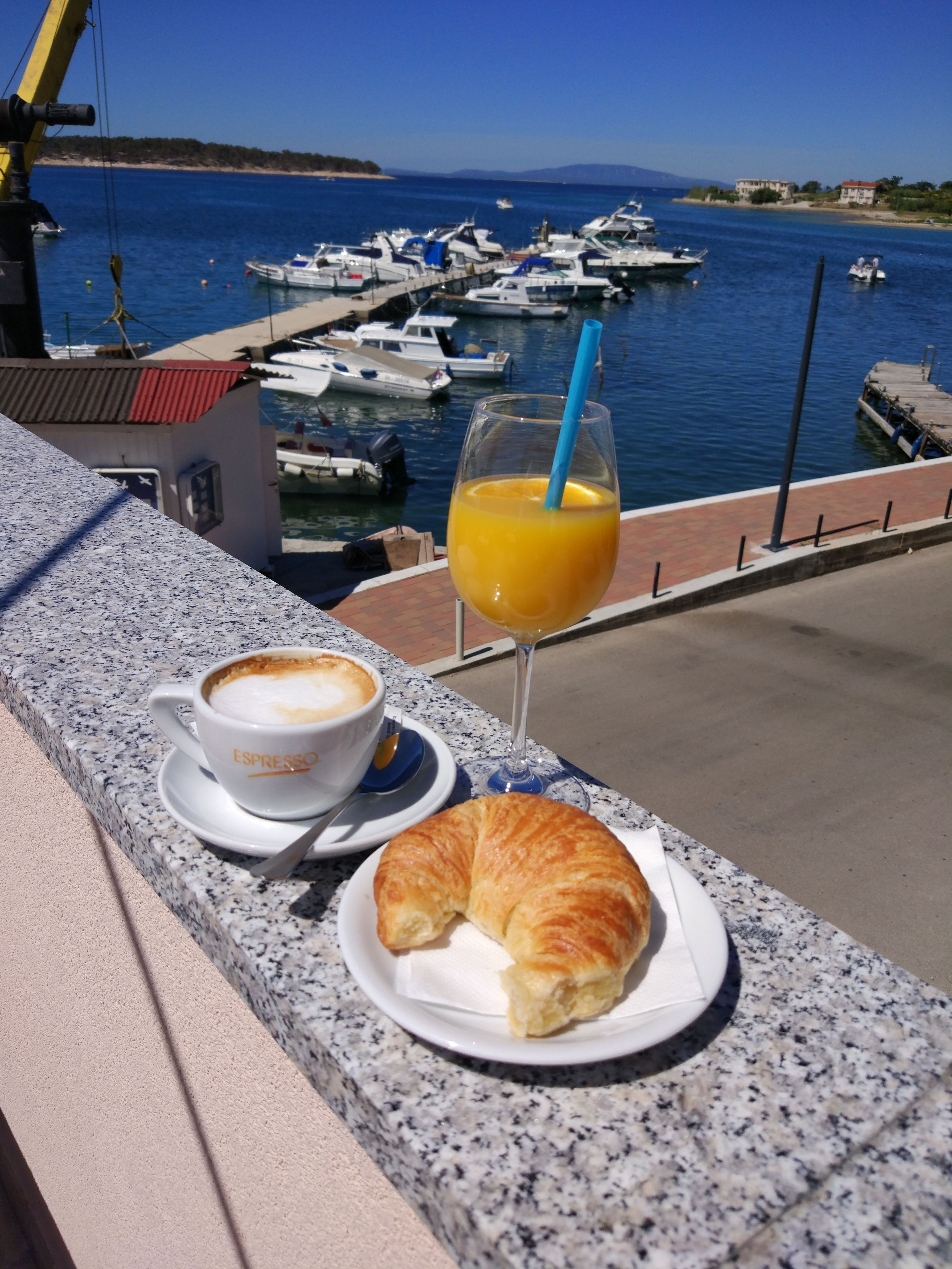 A croissant and espresso on a table with a glass of orange juice, overlooking a marina with boats.