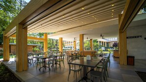 Un área de comedor al aire libre con varias mesas y sillas, vigas de madera en el techo y vista a un edificio al fondo.