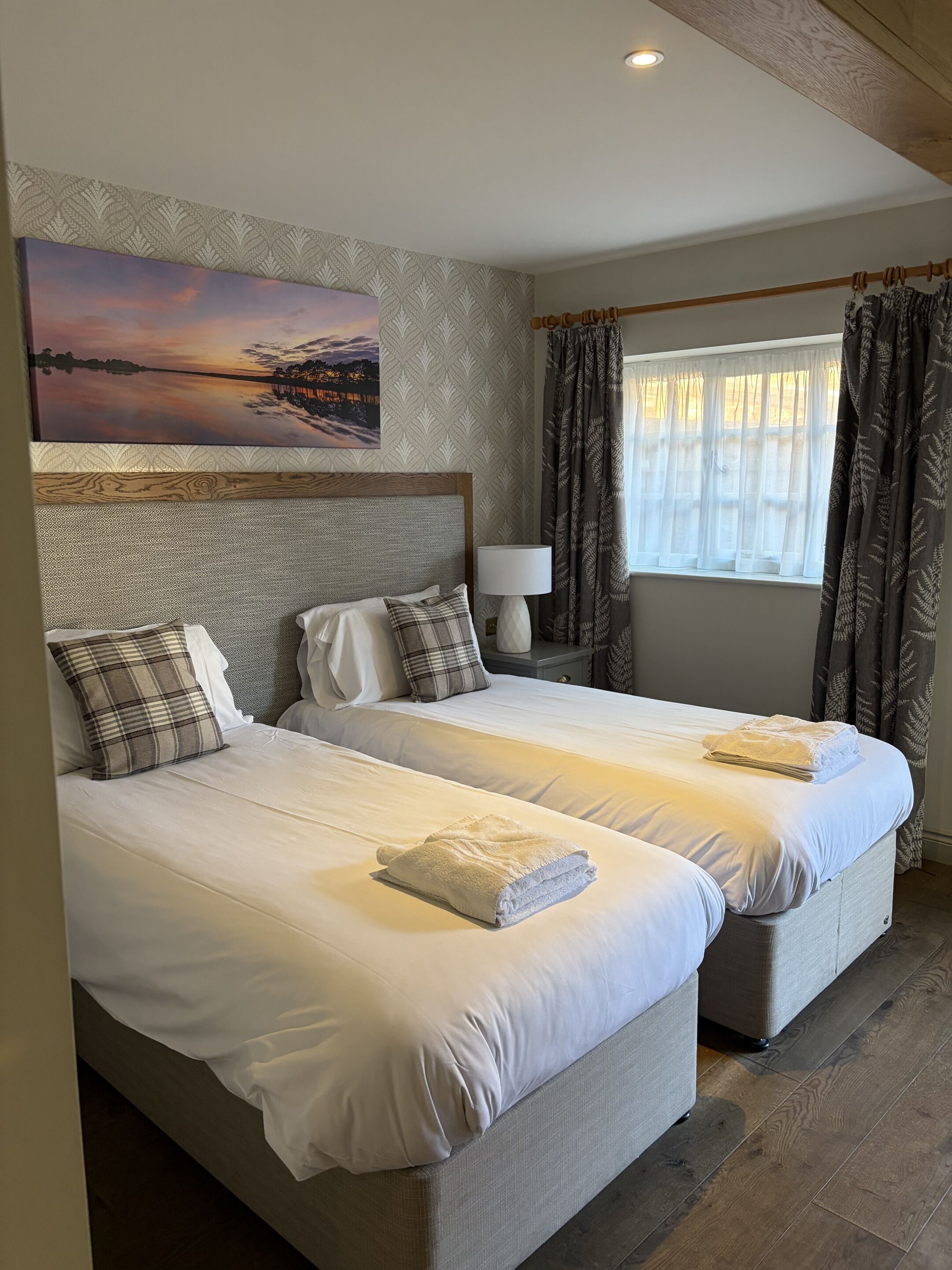 A hotel room with two beds, a painting of a sunset, a window with curtains, and a lamp.