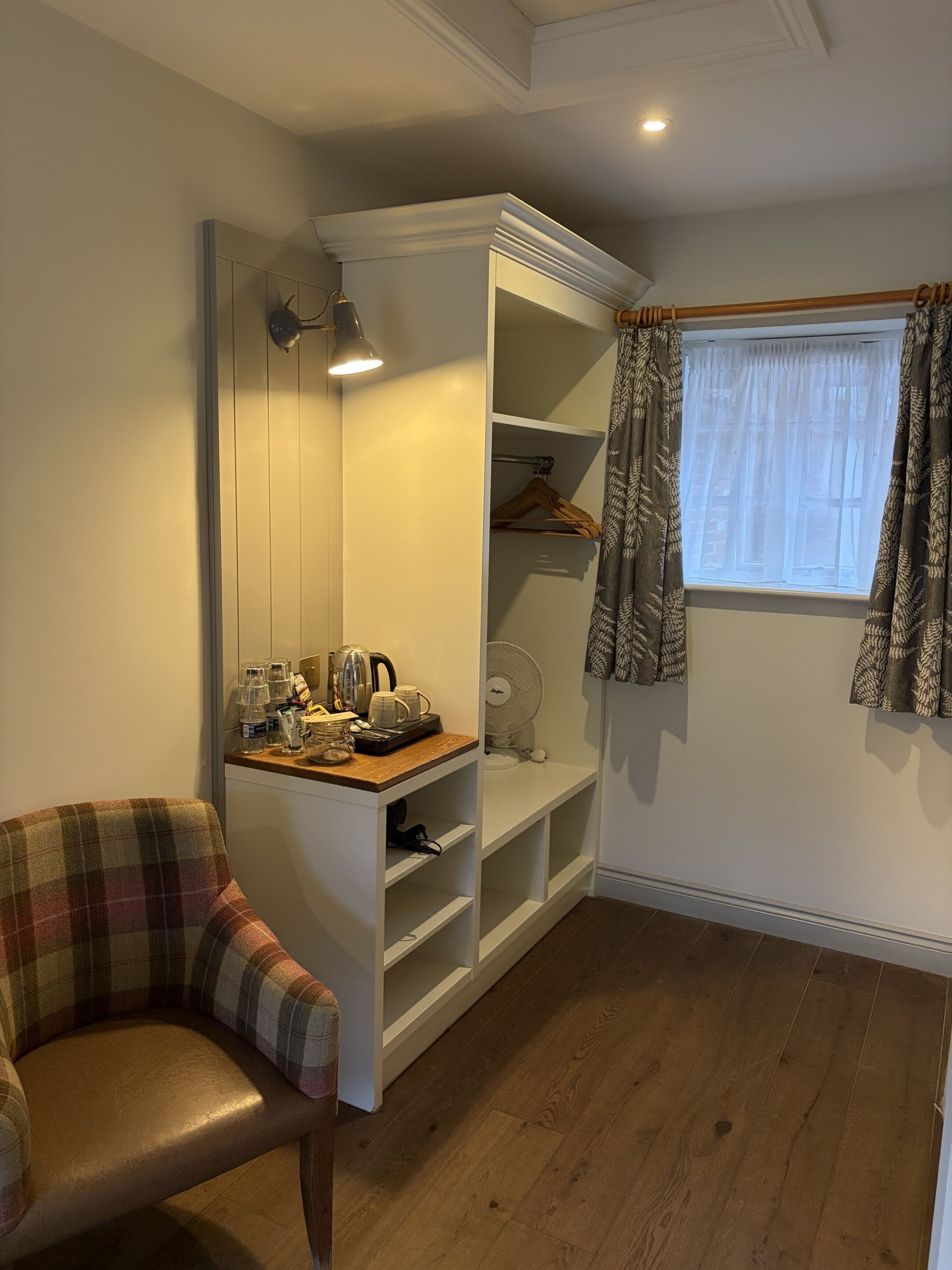 Una habitación acogedora con una butaca escocesa, un estante blanco con un hervidor y una ventana con cortinas estampadas.