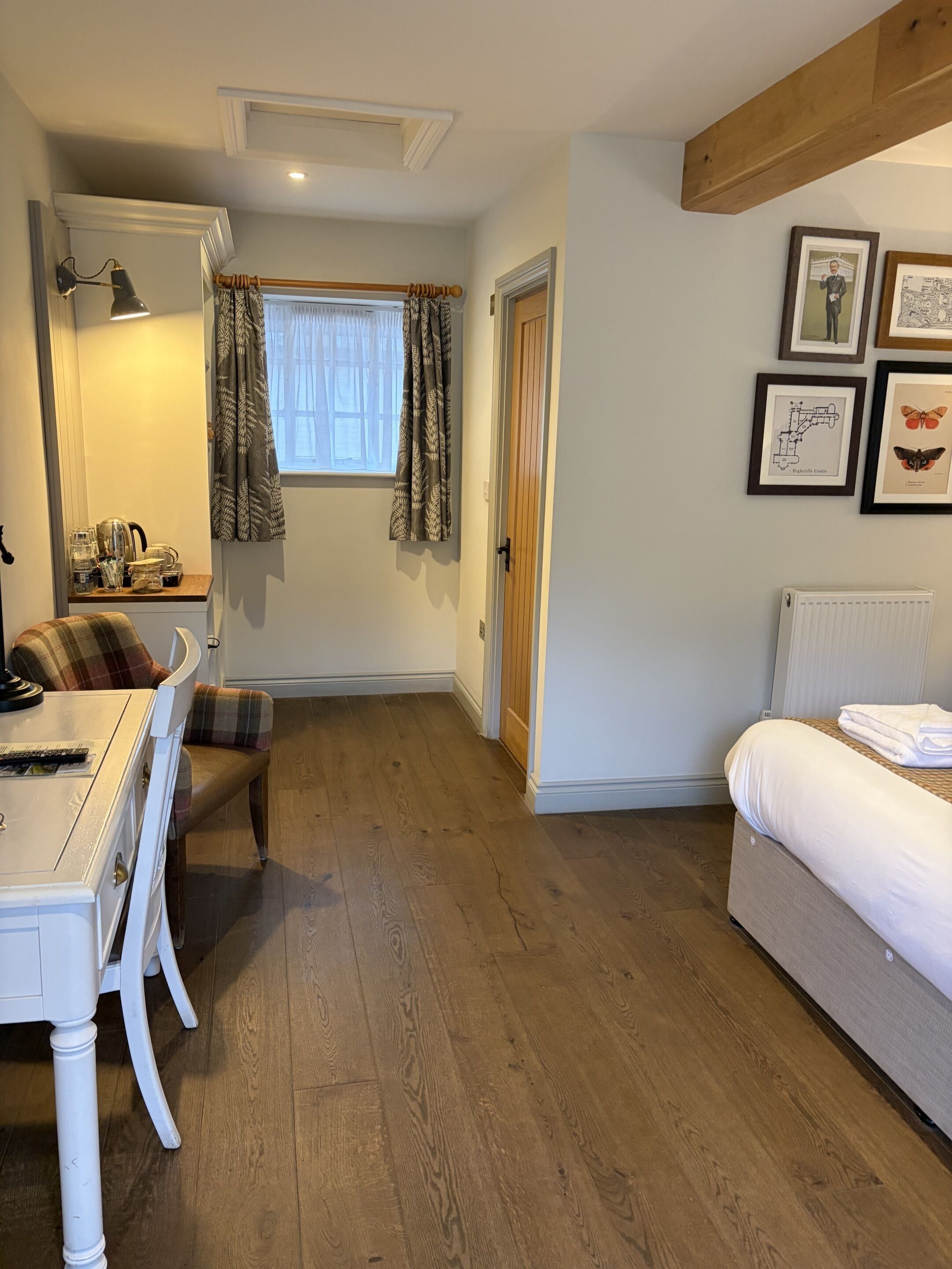 Ein gemütliches Zimmer mit Holzboden, einem weißen Bett, einem Esstisch mit kariertem Stuhl, einer kleinen Küchenzeile mit Wasserkocher und einem Fenster mit gemusterten Vorhängen.