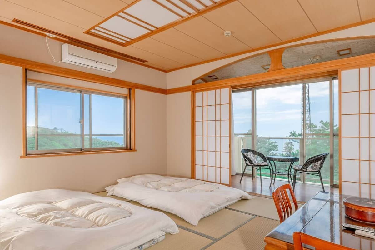 Quarto tradicional japonês com piso de tatame, uma mesa baixa com cadeiras e grandes janelas com vista para o exterior.