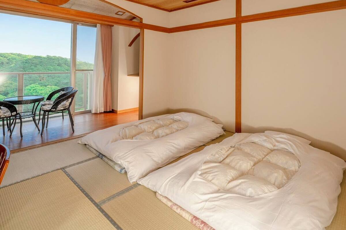 Quarto tradicional japonês com tatames, duas camas, uma varanda com vista e teto de madeira.