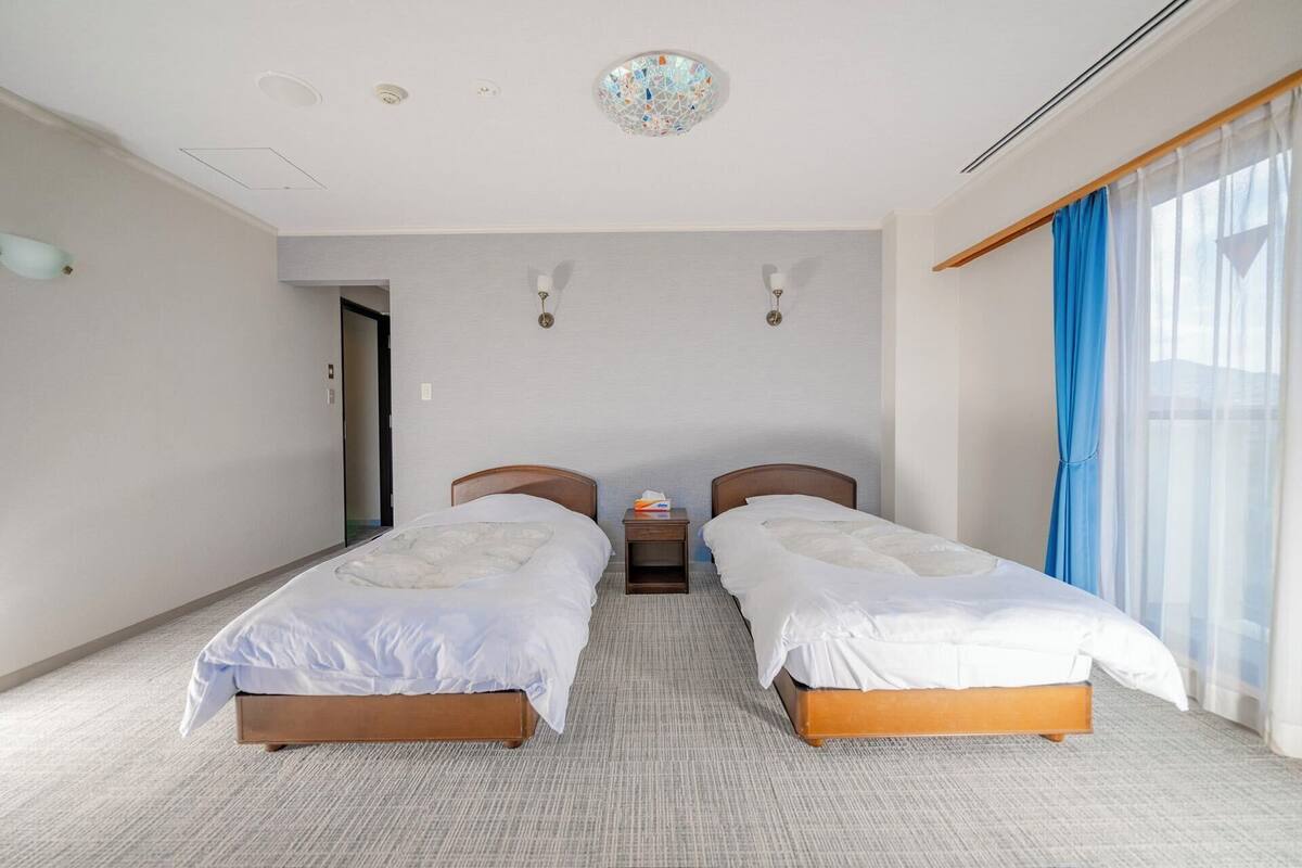 Quarto de hotel com duas camas de solteiro, uma mesinha de cabeceira, luminária no teto e uma janela com cortinas azuis.