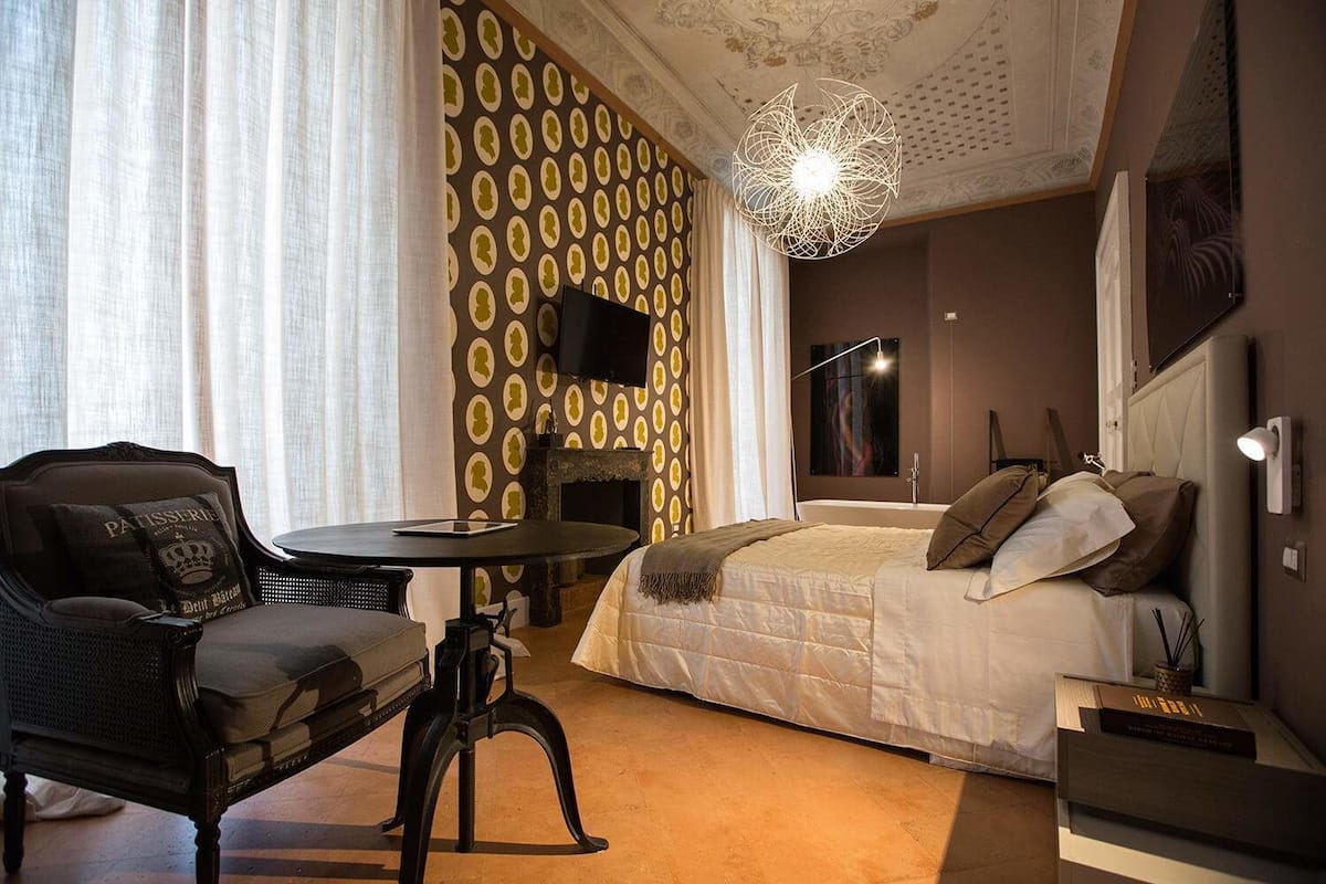 A hotel room with a bed, a chair, a small table, a TV, and a decorative ceiling light.