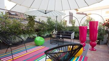 Eine Terrasse im Freien mit einer schwarzen Couch, grünen und gelben Stühlen, einem farbenfrohen Streifen Teppich und einem weißen Sonnenschirm, der Schatten spendet.