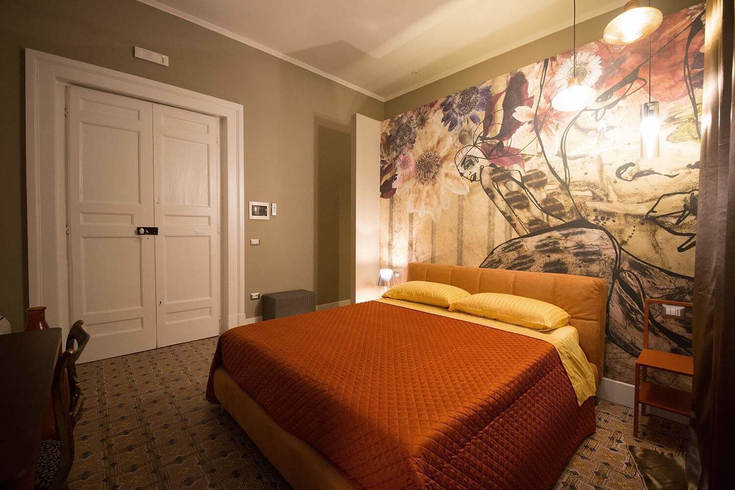 A bedroom with a large bed, a door, a wall with a floral mural, and a small table with a lamp.