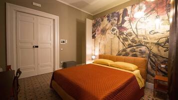 A bedroom with a large bed, a door, a wall with a floral mural, and a small table with a lamp.