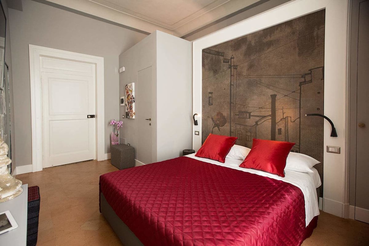 A bedroom with a large bed, a white door, a wall with a cityscape mural, and a small table with a vase of flowers.