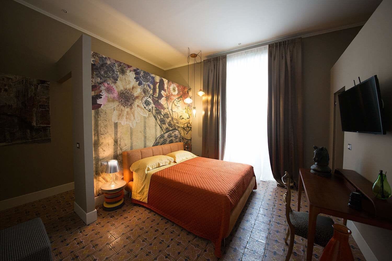 Quarto de hotel com cama, escrivaninha, cadeira, televisão e um grande mural floral na parede.