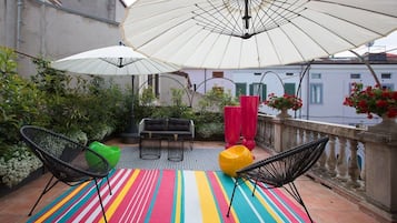 Un balcón con alfombras a rayas, dos sillas negras, un sillón orejero amarillo y sombrillas blancas.